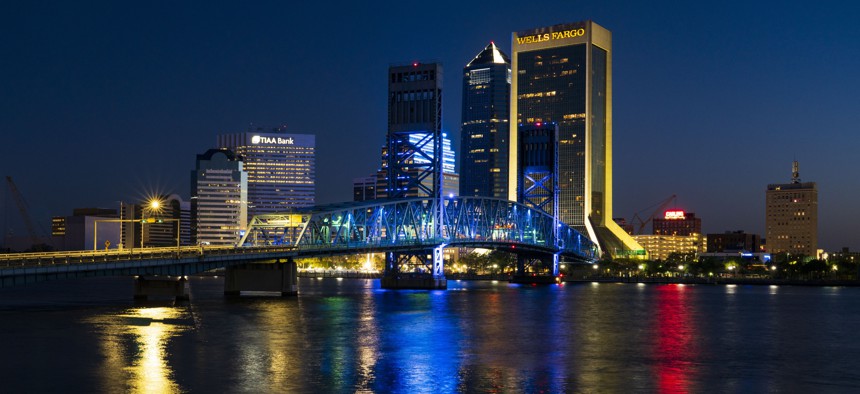Skyline view of Jacksonville features John Alsop Bridge at night with colorful lights reflecting in water. 